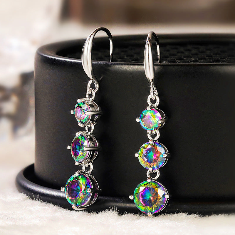 The Prismatic Stellar Studs by Season Prestige are hypoallergenic silver drop earrings with three round iridescent gemstones in a vertical design, shown on a black stand with a soft light background—ideal for any jewelry lover.