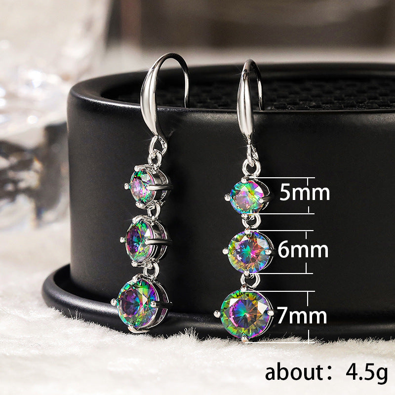 The Prismatic Stellar Studs by Season Prestige are hypoallergenic silver Zirconia earrings, featuring three round, multicolored gemstones (5mm, 6mm, 7mm) and a total weight of ~4.5g, displayed on a sleek black circular stand.