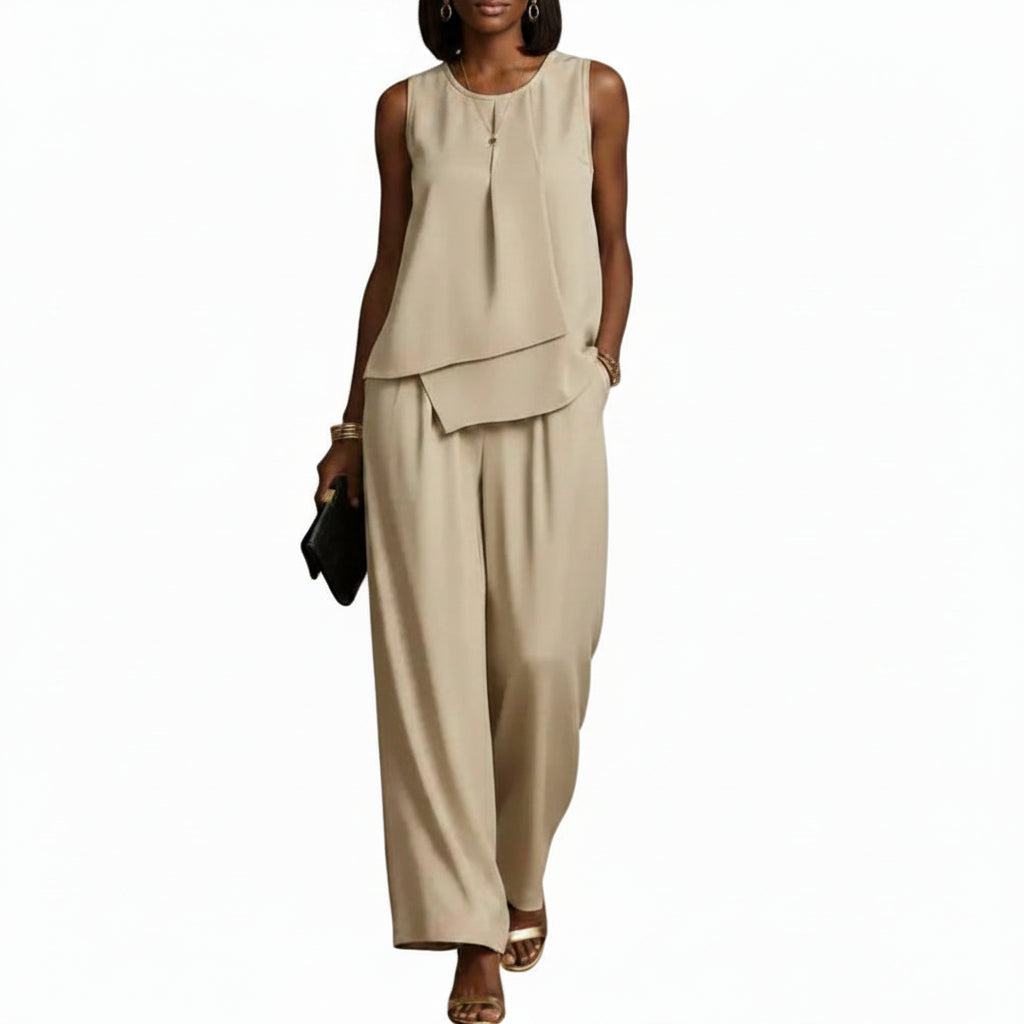 A woman modeling a sleeveless beige two-piece outfit with an asymmetrical draped top and wide-leg pants, holding a small black clutch against a plain white background.