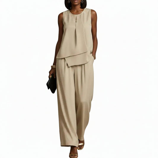 A woman modeling a sleeveless beige two-piece outfit with an asymmetrical draped top and wide-leg pants, holding a small black clutch against a plain white background.