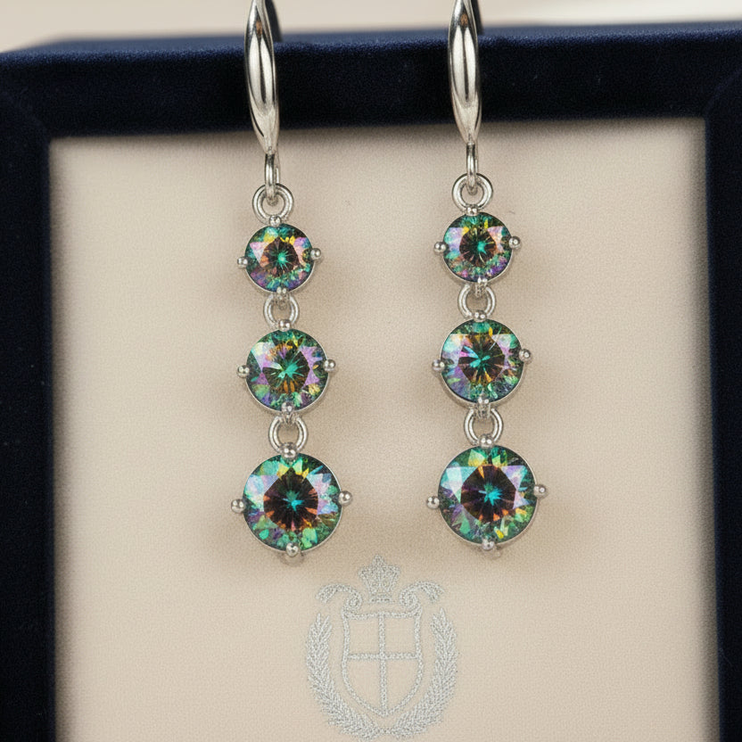 The Prismatic Stellar Studs by Season Prestige are hypoallergenic silver drop earrings with three vertically arranged iridescent gemstones, perfect for sensitive ears and elegantly displayed on a black cylindrical stand.