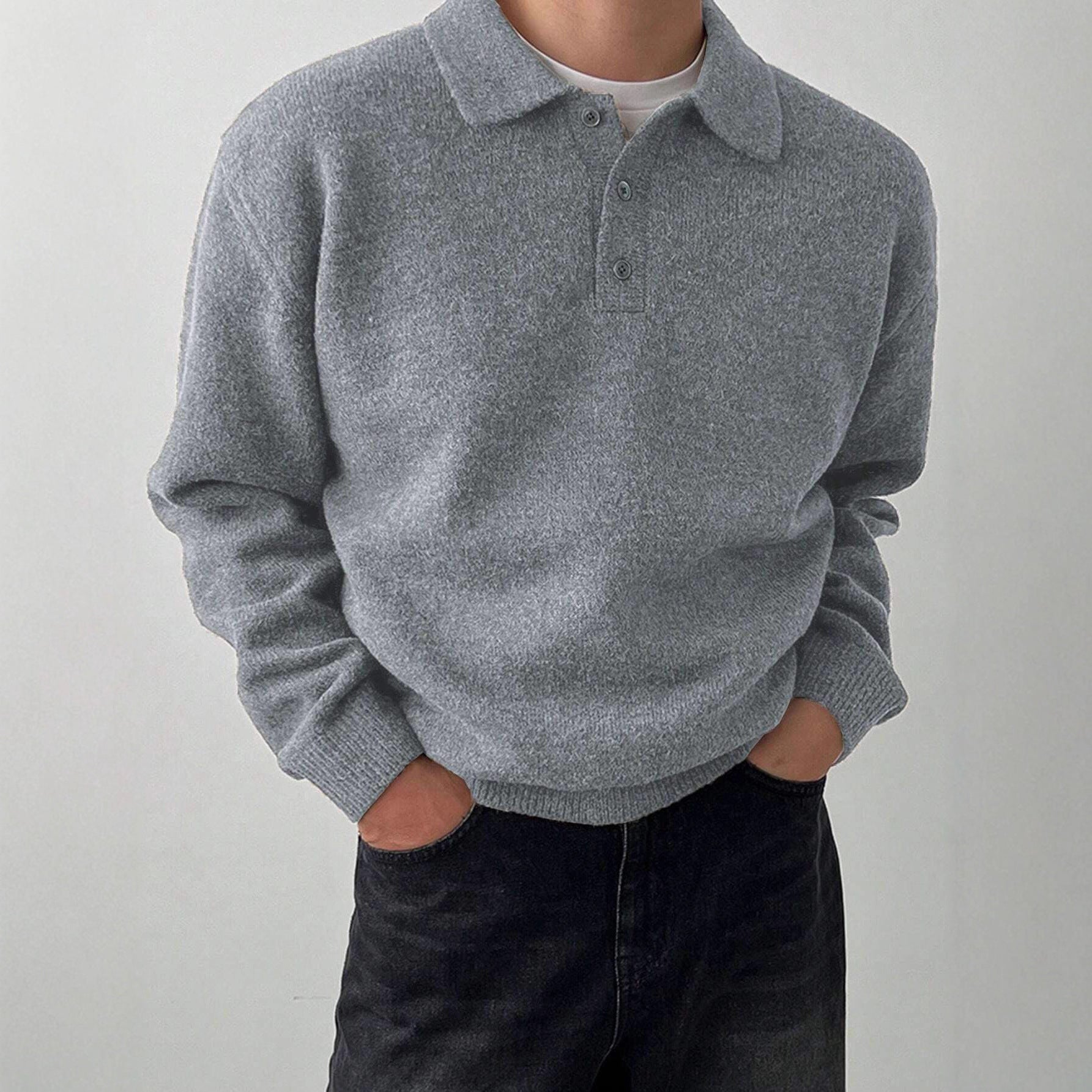 A person wearing a light gray knit polo sweater over a white undershirt and dark jeans, standing against a plain light gray wall.