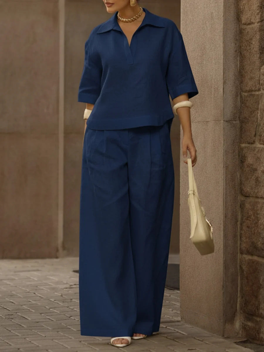 Person wearing a blue outfit with wide-leg pants, holding a beige handbag, standing against a stone wall.