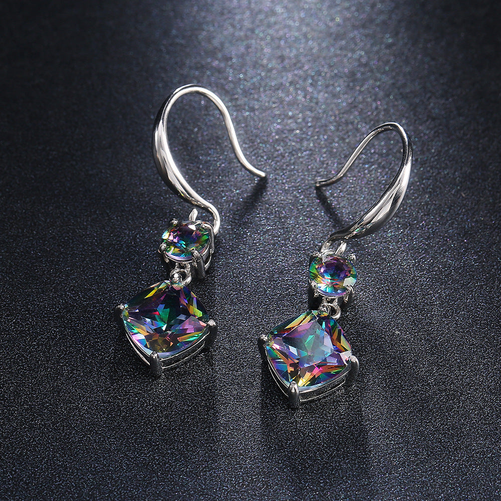 The Prismatic Stellar Studs by Season Prestige are silver dangle earrings with rainbow-colored square and round gemstones, hypoallergenic curved hooks, and sparkling multicolored stones, elegantly displayed on a dark textured surface.