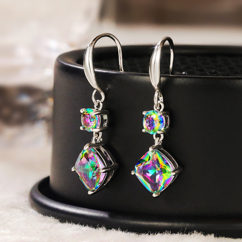 The Prismatic Stellar Studs by Season Prestige are silver dangle earrings with square-cut iridescent gemstones that reflect rainbow colors, creating a vibrant, eye-catching effect; shown on a black circular stand.