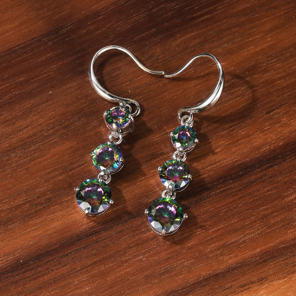 The Prismatic Stellar Studs by Season Prestige are silver drop zirconia earrings, each featuring three round multicolored gemstones, displayed on a polished wooden surface.