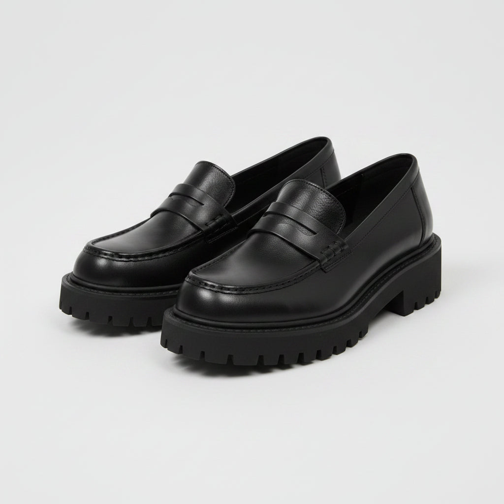 Black platform loafers worn by a person on a light wooden floor with a plant in the background.