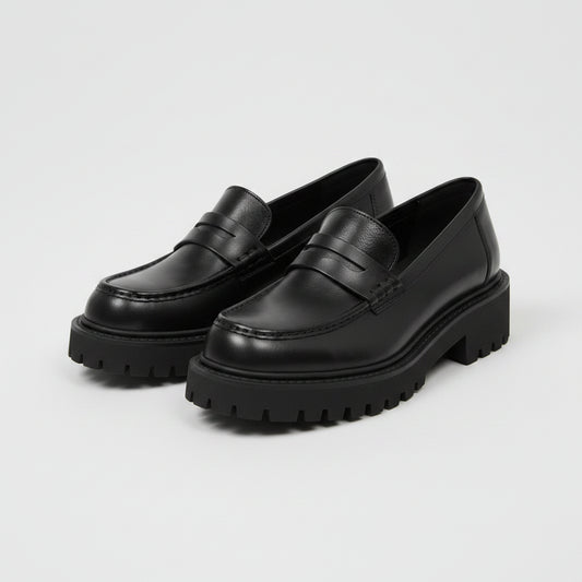 Black platform loafers worn by a person on a light wooden floor with a plant in the background.